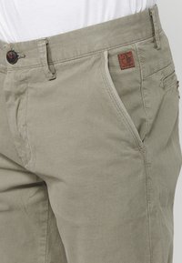 Beige textuurbroek met zijzakken, riemlussen en een bruine vierkante label met een esdoornbladlogo nabij de rechterzak.