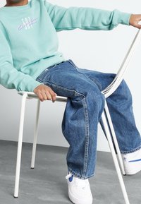 Sudadera verde menta con un gráfico en tonos pastel, jeans de denim azul de corte relajado y zapatillas blancas con detalles azules. Sentado en una silla blanca.