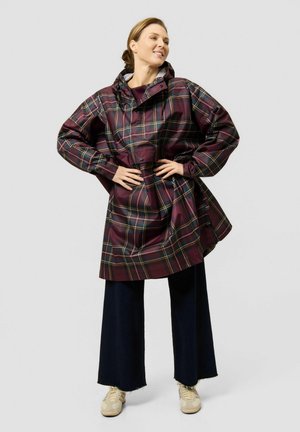 Femme portant un imperméable à carreaux bordeaux avec capuche, pantalon large foncé et baskets beige, debout les mains sur les hanches.