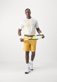 Weißes Sportshirt mit gelben vertikalen Streifen, kombiniert mit orangefarbenen Shorts. Trägt weiße Tennisschuhe und Handgelenkbänder, hält einen Tennisschläger.