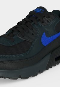 Κοντινό πλάνο μαύρου και σκούρου πράσινου sneaker Nike με μαύρα κορδόνια και μπλε λογότυπο Nike swoosh στο πλάι.