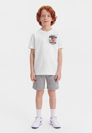 Barn med rødt hår iført en hvid T-shirt med grafik, grå shorts, hvide sokker og hvide sneakers med lilla accenter.