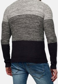 Man som bär en svart och grå stickad tröja med färgblock och blå jeans, står med ryggen mot betraktaren.