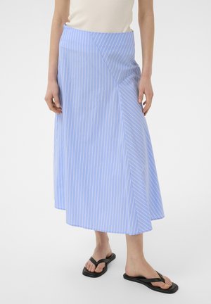 Kobieta w jasnoniebieskiej i białej spódnicy midi w paski z asymetrycznym wzorem i czarnych japonkach.