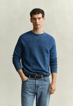 Junger Mann steht mit einer Hand in der Tasche, trägt ein blaues Sweatshirt, einen geflochtenen dunklen Gürtel und hellblaue Jeans vor einfachem Hintergrund.