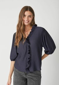 Blusa blu navy in tessuto texturizzato e stropicciato, con scollatura a V arricciata e maniche a sbuffo, abbinata a jeans grigi.