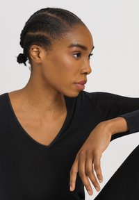 Top nero a maniche lunghe con scollatura a V, caratterizzato da una texture liscia e un bordo sottile. Il modello ha i capelli intrecciati, mostrando il profilo laterale e la posizione della mano.