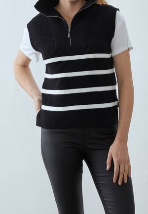 Personne portant un gilet en tricot noir sans manches avec des rayures horizontales blanches et une fermeture éclair sur une chemise blanche à manches courtes et un pantalon noir.