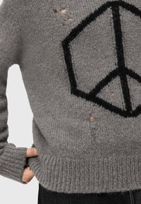 Maglione grigio lavorato a maglia con simbolo di pace geometrico nero e buchi di usura intenzionali, indossato con una mano infilata nella tasca laterale.