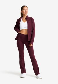 Mörkröd idrottsjacka med dragkedja, kombinerad med en vit crop top och matchande mörkröda leggings. Vita skor fulländar looken. Slätt tyg.