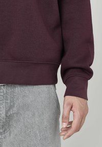 Sweatshirt bordeaux avec des poignets et un ourlet côtelés, fabriqué en tissu doux en mélange de coton, associé à un pantalon gris texturé. Main visible sur le côté.