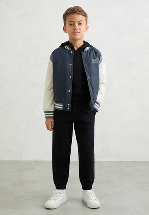 Laivastonsininen ja kerma varsity-takki raidoitetuilla resoreilla, jossa on painonapit ja kirjailtu logo. Päällä mustan hupun ja mustien housujen kanssa.