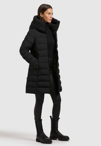 Svart vadderad lång kappa med hög krage, med dragkedja framtill och sidofickor. Bärs över svarta tajta leggings och svarta ankelboots.