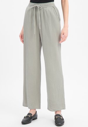 Pantalon large en tissu gris clair froissé avec taille à cordon, porté avec des mocassins noirs et un haut noir, les mains reposant le long du corps.