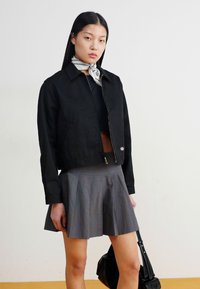 Veste noire cintrée avec fermeture éclair à l'avant, associée à une jupe plissée grise. Les accessoires notables incluent un sac noir et une écharpe blanche à pois.