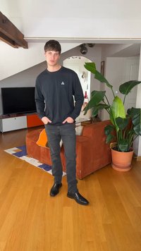 Ung mann i svart langermet skjorte og jeans står med hendene i lommene i en moderne stue ved siden av en stor potteplante og en oransje sofa.