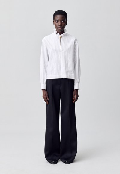 Persona con una blusa de cuello rizado blanco con un broche dorado y pantalones negros de pierna ancha, de pie contra un fondo claro y liso.