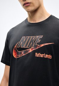 Άνδρας που φοράει μαύρο t-shirt Nike με μεγάλο πορτοκαλί λογότυπο Swoosh και την επιγραφή «Netherlands», εμφανιζόμενος από τον λαιμό μέχρι τη μέση του κορμού.