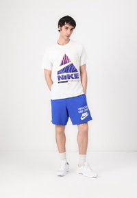 Nike grafiska t-kreklu krēmkrāsā ar purpura, rozā un zaļo dizainu; kombinēts ar zilām šortiem, kas rotāti ar baltu logo un tekstu. Baltas kedas.