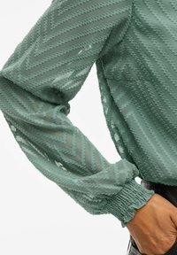 Blusa testurizzata verde menta con maniche a sbuffo, motivo a zigzag e polsini elasticizzati. Il tessuto trasparente aggiunge profondità e dimensione.