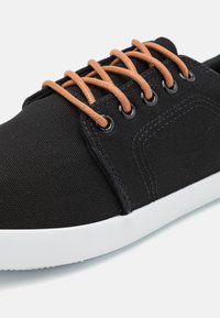 Zapatilla de lona negra con cordones marrones. Presenta ojales redondos negros y una suela de goma blanca. Se puede apreciar un tejido texturizado y detalles cosidos.