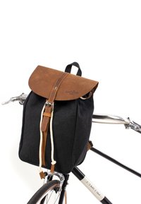 Zaino in tela nera con una patta in pelle marrone, dotato di chiusura con cordino, accentato in pelle e maniglia superiore, fissato a una bicicletta.