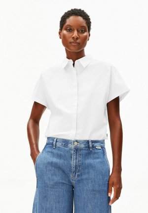 Frau trägt ein kurzärmeliges weißes Hemd mit Kragen und hoch taillierte blaue Jeans, steht mit einer Hand in der Tasche.