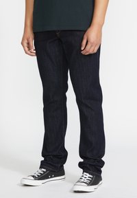 Jean en denim foncé coupe slim, avec une texture subtile et des coutures orange contrastantes. Porté avec des baskets noires et des lacets blancs.
