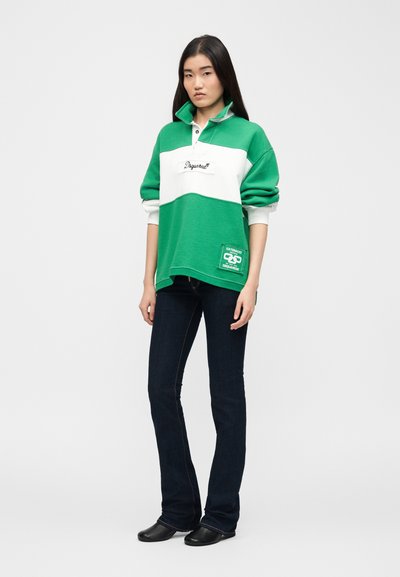 Νεαρή γυναίκα με μακριά σκούρα μαλλιά, φορώντας πράσινο και λευκό φούτερ, σκούρα μπλε τζιν και μαύρα παπούτσια, στέκεται μπροστά σε απλό φόντο.