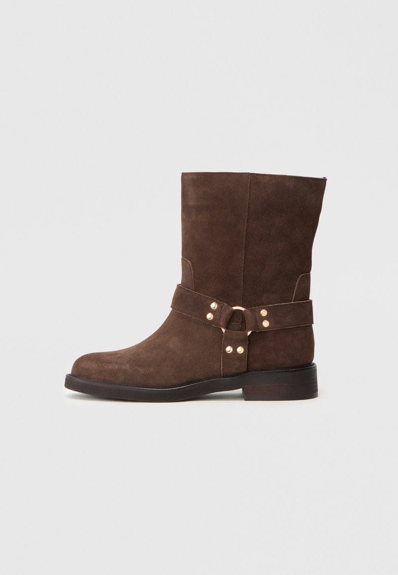 Braune Wildlederschuhe mit rundem Zehenbereich, ausgestattet mit einem breiten Riemen und metallischen Akzenten. Die Sohle ist aus dunklem Holz und hat einen leichten Absatz.