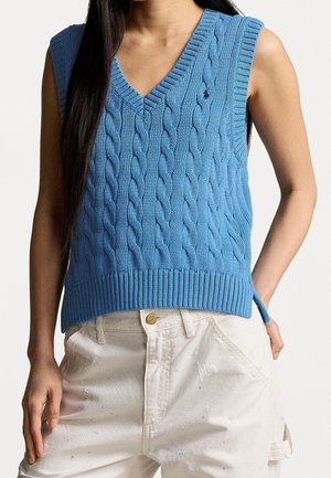 Blau gestrickte ärmellose V-Ausschnitt-Weste mit Zopfmuster, getragen mit offwhite Hosen mit Farbspritzerdetails.