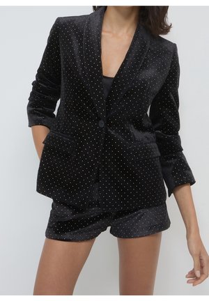 Nainen pukeutuneena mustaan samettiseen bleiseriin ja siihen sopiviin lyhyisiin housuihin, joissa on kultaiset pilkut, poseeraa rentoina käsinä sivuilla.