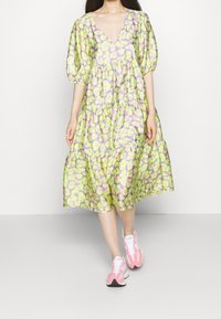 Šaty s výstřihem do V a nabíranými rukávy v zelené barvě, s fialovým a žlutým květinovým vzorem, s vrstvenou sukní, doplněné o růžové tenisky. Hladká textura látky.