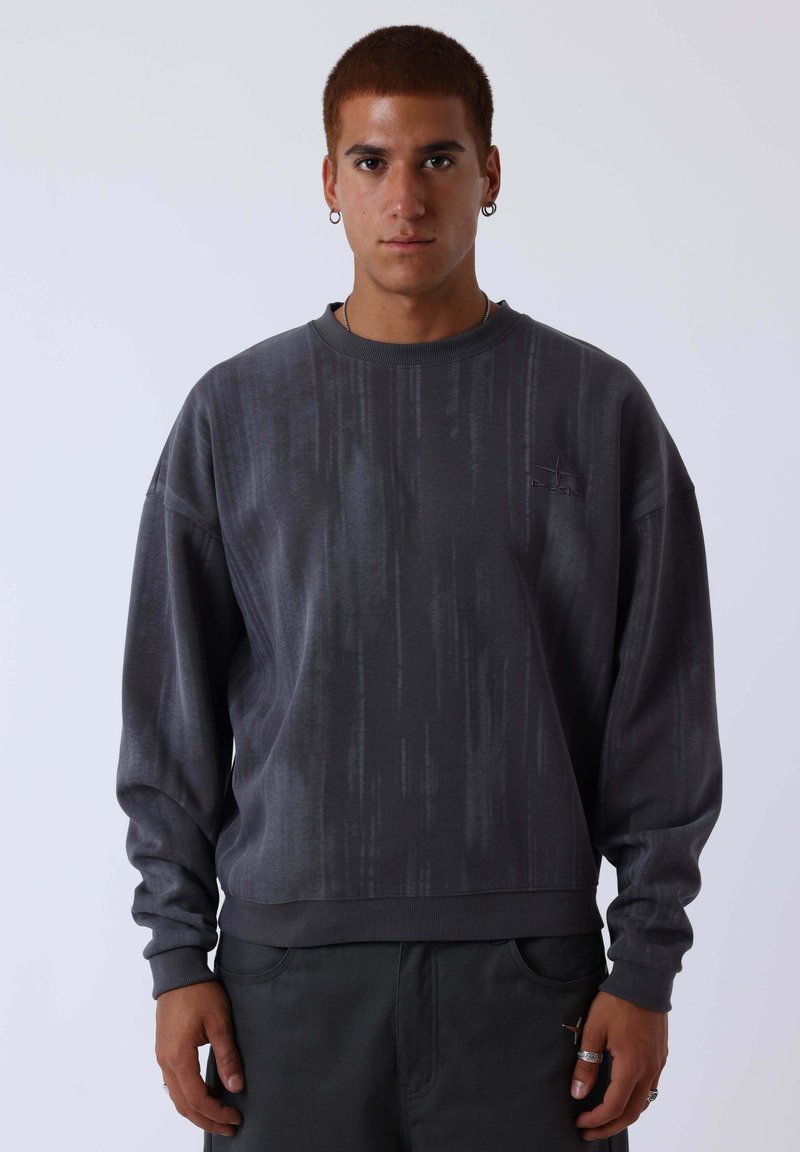 Jeune homme aux cheveux roux courts portant un sweat-shirt texturé gris foncé et un pantalon foncé, debout devant un fond clair uni.