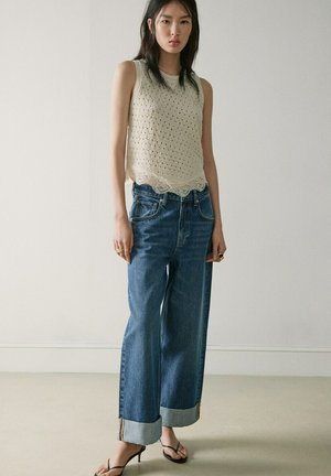 Jeune femme debout, portant un haut en maille crème sans manches, un jean bleu large avec poignets retroussés, des sandales à talons noires et des bagues en or, devant un mur uni.