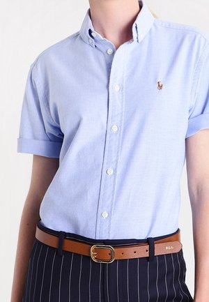 Kobieta ubrana w jasnoniebieską koszulę z krótkim rękawem zapinaną na guziki, z podwiniętymi mankietami, brązowy skórzany pasek i ciemne spodnie w prążki.