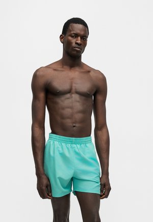 Uomo muscoloso in piedi contro uno sfondo bianco, indossa pantaloncini sportivi turchesi con le mani rilassate lungo i fianchi.