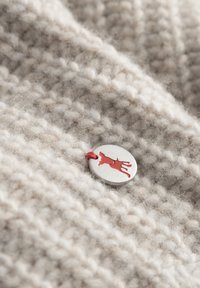 Beige wollen stof met een gestructureerde brei, met een metalen knop met een rood dierenontwerp bevestigd met een rode draad.