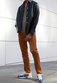 Czarna skórzana kurtka-koszula, beżowa prążkowana koszula, brązowe spodnie oraz niebieskie buty sportowe z żółtymi akcentami. Gładka tekstura i dopasowany krój.