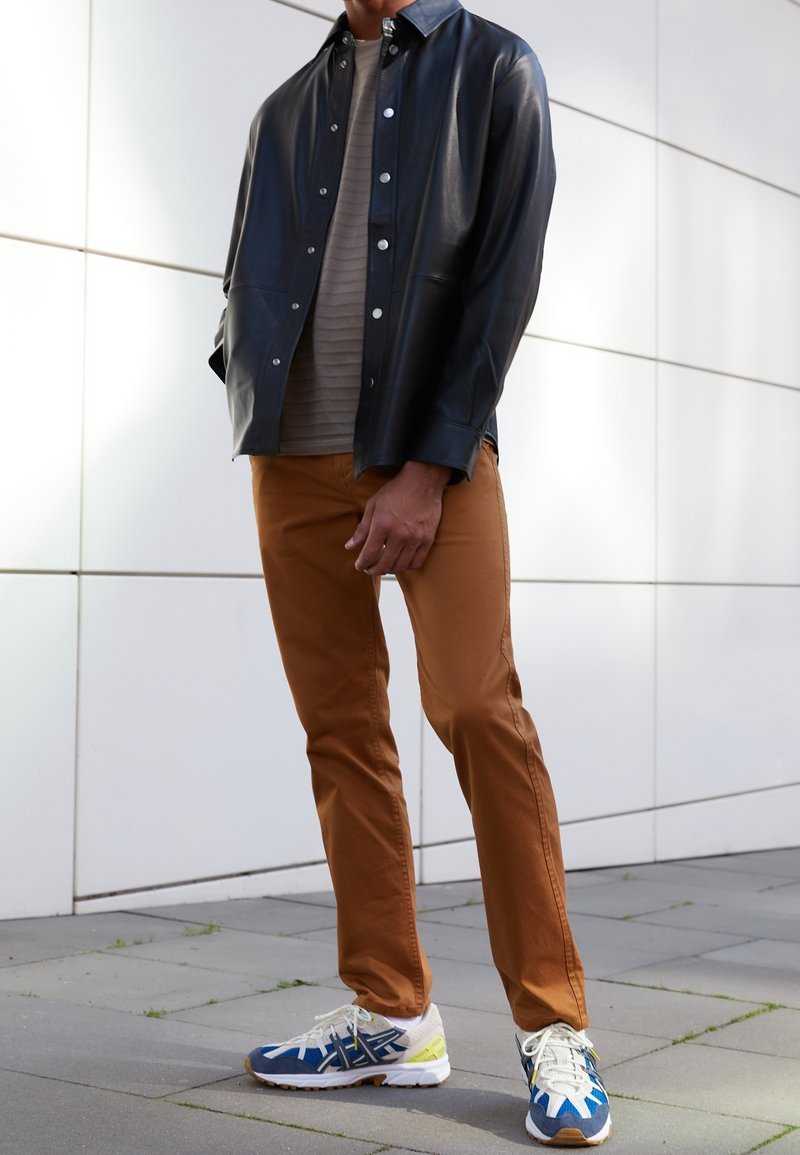 Czarna skórzana kurtka-koszula, beżowa prążkowana koszula, brązowe spodnie oraz niebieskie buty sportowe z żółtymi akcentami. Gładka tekstura i dopasowany krój.