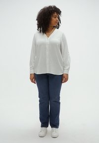 Άσπρο, υφασμάτινο πουκάμισο με κουμπιά, λαιμόκοψη σε σχήμα V και μακριά μανίκια, συνδυασμένο με σκούρο μπλε τζιν παντελόνι με ίσια μπατζάκια και άσπρα αθλητικά παπούτσια.