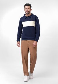 Man walking wearing a navy blue and white striped long sleeve shirt, brown pants, and white sneakers on a plain white background.