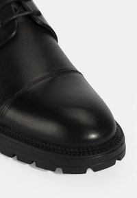 Zwarte leren schoen met een ronde neus, matte en glanzende texturen, gestikte details en een robuuste rubberen zool met gripgroeven.