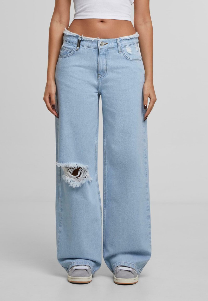 Svetlomodré džínsy so širokými nohavicami a rozstrapkaným pásom, s veľkou poškodenou dierou na ľavnom kolene. Vyrobené z denimového materiálu, s striebornými prvkami.