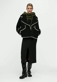 Frau trägt eine schwarze Wolljacke mit weißer Naht, einen grünen Kapuzenpullover, einen schwarzen Midirock mit Schlitz vorne und schwarze kniehohe Stiefel.