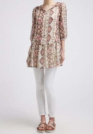 Tunique beige avec un motif géométrique dans des nuances de rose et de marron, dotée d'un décolleté en arrondi, de manches trois-quarts et d'un ourlet en couches. Associée à des leggings blancs et des sandales métalliques.