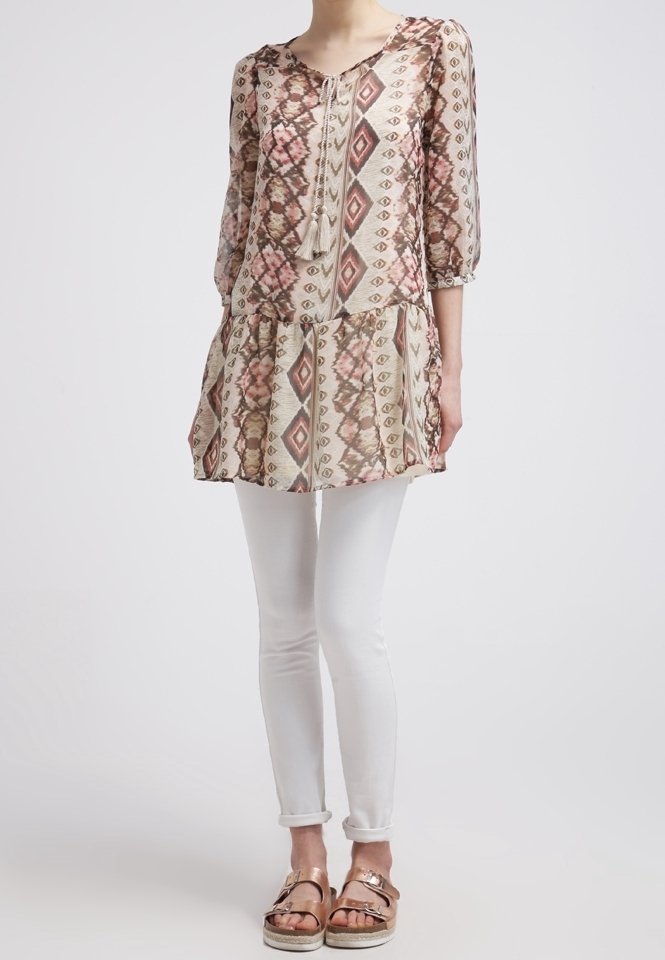 Túnica beige con un patrón geométrico en tonos de rosa y marrón, con escote en forma de barco, mangas de tres cuartos y un dobladillo en capas. Combinada con leggings blancos y sandalias metálicas.