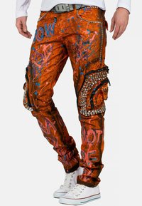 Oranje jeans met spetterverf in blauw en roze. Beschikt over ritszakken en metalen studs aan de zijkant. Rechte pasvorm.