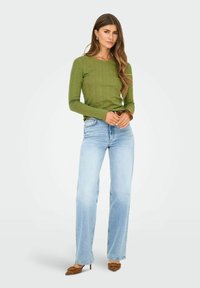 Groene geribbelde langemouwen top, lichtblauwe rechte jeans en bruine spitsneus hakken. Model staat met handen gevouwen, neutrale achtergrond.