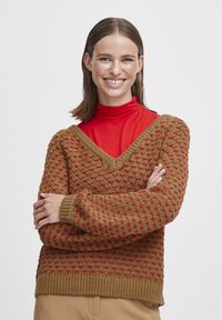 Pull en tricot brun, avec un col en V et un motif texturé avec des accents rouges. Manches et ourlet côtelés, porté par-dessus un col roulé rouge.